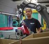 Ein Roboterarm hebt ein Plastikteil vom Fließband. Im Hintergrund ist eine Angestellte des Unternehmens zu erkennen.