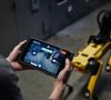Zwei Hände halten ein Tablet, über das der Roboterhund Spot von Boston Dynamics im Hintergrund ferngesteuert wird
